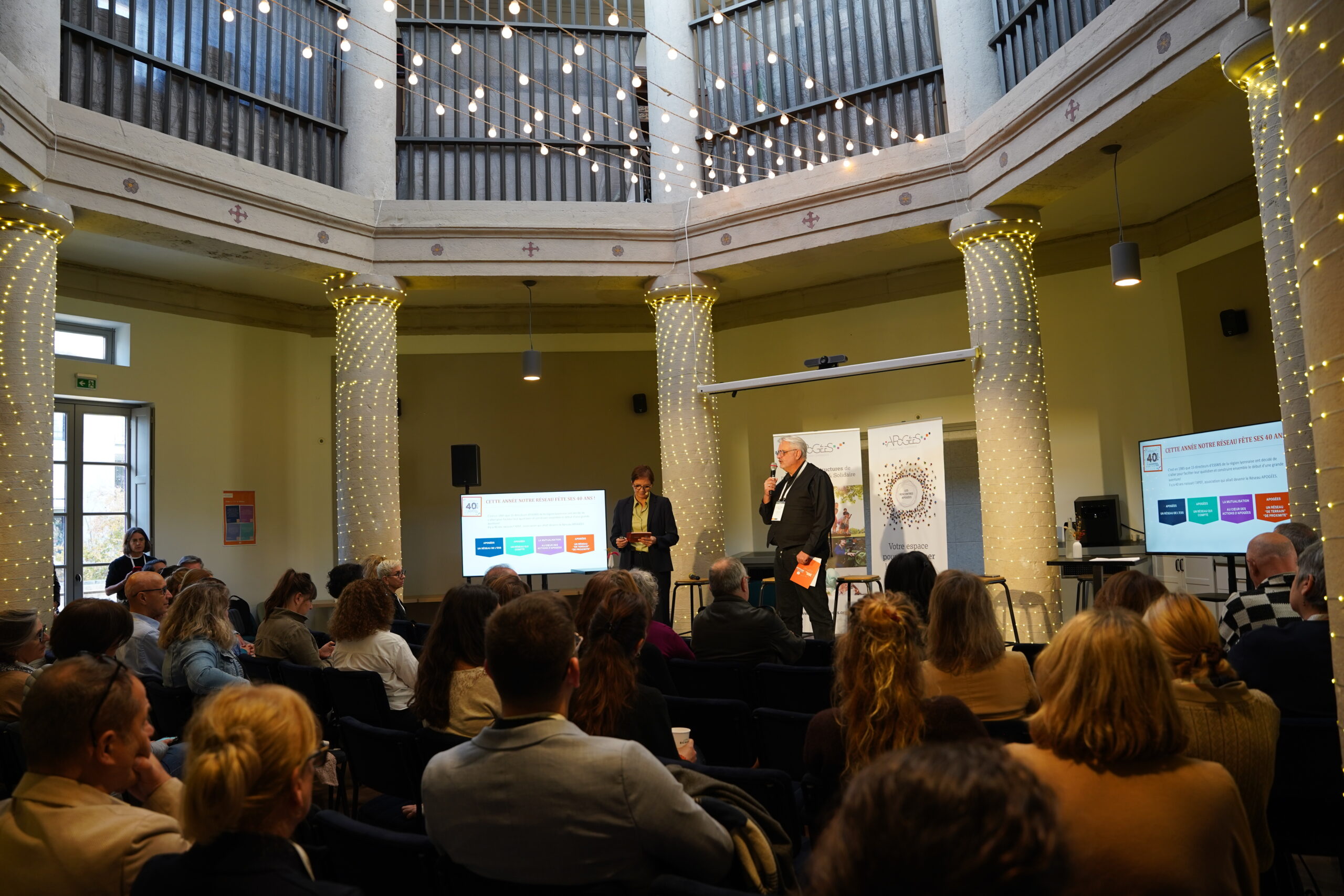Conférence APOGÉES avec intervenants et public dans une salle décorée de guirlandes lumineuses